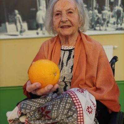 Sala. Seniorka siedzi na wózku. Uśmiecha się. Nogi ma okryte kocem. Wyciąga rękę, w dłoni trzyma pomarańczę.
