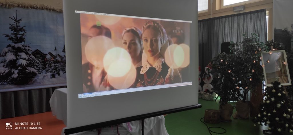 Sala. Choinka, świąteczne dekoracje. Na ekranie projektora zbliżenie dwóch kobiet i mężczyzny w strojach ludowych.