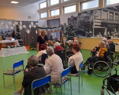Sala. Świąteczne dekoracje. Seniorzy siedzą w rzędach. Dyrektorka Agnieszka Cysewska pokazuje światełko Betlejemskie.
