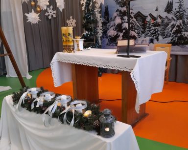 Sala. Świąteczne dekoracje. Przed ołtarzem stół. Na nim stroik z gałązek choinki. Obok światełko Betlejemskie.