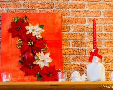 Na półce świąteczny obraz z bombek i gwiazd betlejemskich. Obok świeczniki, figurka aniołka, świeczka.