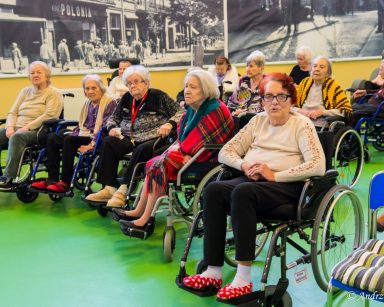 Sala. Seniorzy siedzą w rzędach. Część siedzi na wózkach inwalidzkich, inni na krzesłach.