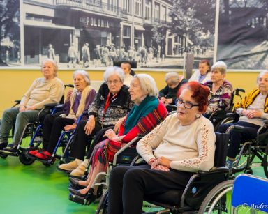 Sala. Seniorzy i pracownicy siedzą w rzędach. Część siedzi na wózkach inwalidzkich, inni na krzesłach.
