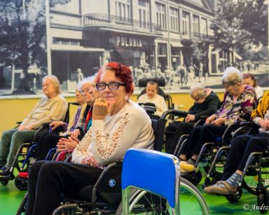 Sala. Seniorzy siedzą w rzędach. Część siedzi na wózkach inwalidzkich, inni na krzesłach.