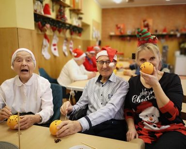 Sala ze świątecznymi ozdobami. Przy stołach seniorzy, terapeutka Beata Gadomska. Dekorują pomarańcze goździkami.