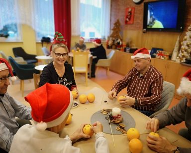 Sala ze świątecznymi ozdobami. Przy stołach seniorzy, terapeutka Beata Gadomska. Dekorują pomarańcze goździkami.