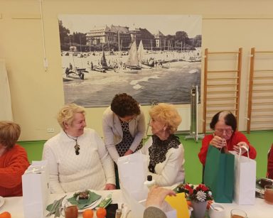 Sala. Seniorzy przy stole. Wiceprezydentka Magdalena Czarzyńska-Jachim rozdaje prezenty, rozmawia z seniorami.
