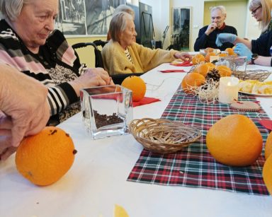 Sala. Przy stole dyrektorka Agnieszka Cysewska, seniorzy. Dekorują pomarańcze goździkami.