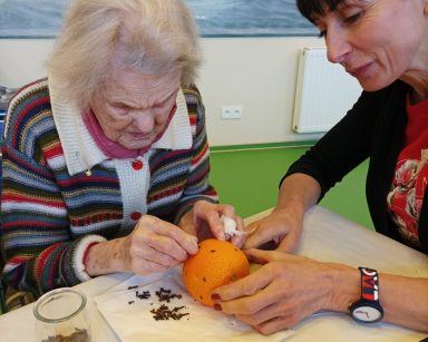 Sala. Przy stole terapeutka Ania Rzepczyńska i seniorka. Wspólnie dekorują pomarańczę goździkami.