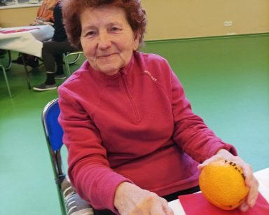 Sala. Seniorka dekoruje pomarańczę goździkami. Uśmiecha się. W tle pracownicy i seniorzy przy drugim stole.