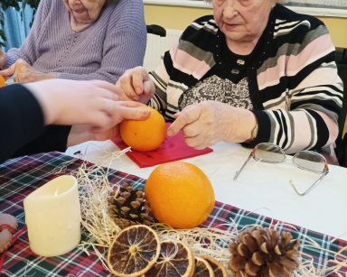 Przy stole siedzą seniorki. Dekorują pomarańcze goździki. Na stole świąteczna dekoracja, szyszki, suszone pomarańcze.