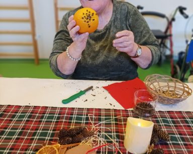 Przy stole seniorka. Śmieje się i pokazuje pomarańczę ozdobioną goździkami. Na stole świąteczna dekoracja, szyszki, pierniki.