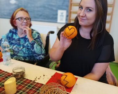 Przy stole fizjoterapeutka Paulina Winczura. Trzyma pomarańczę z goździkami wbitymi w kształt serca. Obok seniorka.