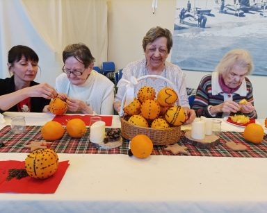 Przy stole seniorki, terapeutka Ania Rzepczyńska. Dekorują pomarańcze goździkami. Na stole kosz z ozdobionymi pomarańczami.