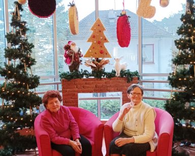 Dwie seniorki w fotelach. Na tle okna świąteczna dekoracja, dwie choinki, kominek, ozdoby ze styropianu na wstążkach.