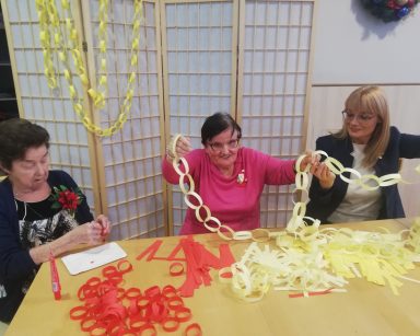 Sala. Przy stole dyrektorka Agnieszka Cysewska, dwie seniorki. Wspólnie robią papierowe łańcuchy.