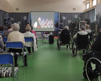 Duża sala. Świąteczne dekoracje, choinki z bombkami. Seniorzy siedzą w rzędach przed ekranem projektora. Oglądają koncert.