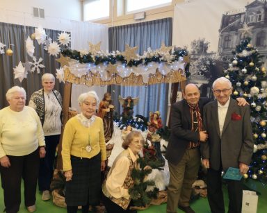 Sala. Świąteczne dekoracje. Seniorzy pozują do zdjęcia przy szopce. obok stoi udekorowana choinka.