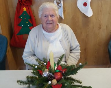 Sala. Świąteczne dekoracje. Seniorka siedzi przy stole. Na blacie świąteczny stroik z gałązek choinki, bombek i świeczki.