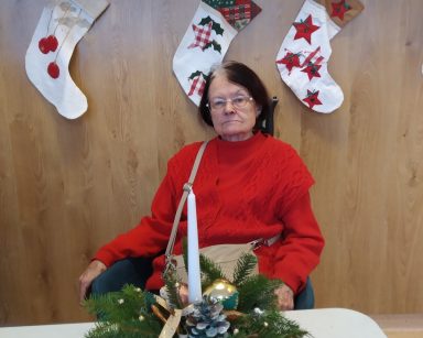 Sala. Świąteczne dekoracje. Seniorka siedzi przy stole. Na blacie świąteczny stroik z gałązek choinki, bombek i świeczki.