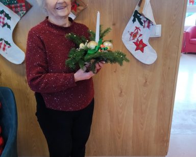 Sala. Świąteczne dekoracje. Seniorka stoi. Trzyma w dłoniach świąteczny stroik z gałązek choinki, bombek i świeczki.