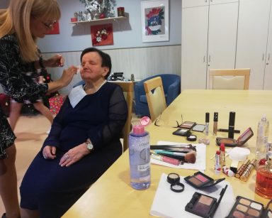 Świetlica. Na blacie stołu kosmetyki. Dyrektorka Agnieszka Cysewska robi makijaż seniorce.