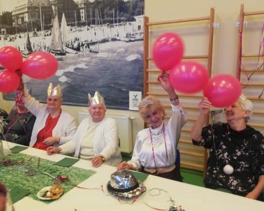 Sala. Przy stole seniorzy. Mają na głowach korony. Machają różowymi balonami.