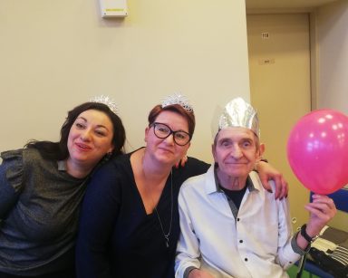 Senior ma srebrną koronę, w ręku balon. Obok terapeutka Gosia Jancelewicz i koordynatorka Edyta Życzyńska. Śmieją się.