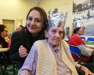Sala. Seniorka w srebrnej koronie. Obejmuje ją fizjoterapeutka Paulina Winczura. Przy stołach siedzą seniorzy.