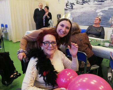 Sala. Seniorka trzyma balony śmieje się. Za nią fizjoterapeutka Martyna Józefczyk. Przy stołach seniorzy.