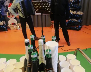 Sala. Przy mikrofonie kierownik Arkadiusz Wanat, obok muzyk z akordeonem. Przed nimi kubki, butelki szampana.