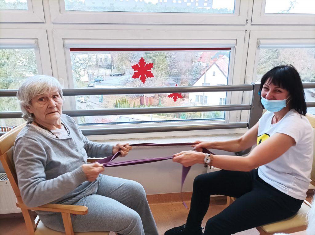 Przy oknie, na przeciwko siebie siedzą terapeutka Ania Rzepczyńska i seniorka. Ćwiczą wspólnie, rozciągają taśmę.