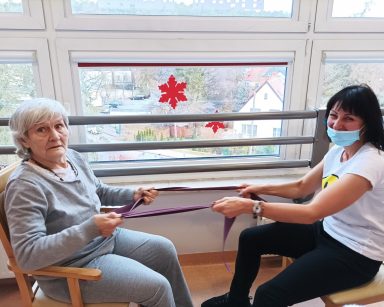 Przy oknie, na przeciwko siebie siedzą terapeutka Ania Rzepczyńska i seniorka. Ćwiczą wspólnie, rozciągają taśmę.