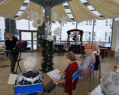 Ogród zimowy. Terapeutka Beata Brzozowska stoi przy pianinie. Gra i śpiewa. Przed nią na krzesłach seniorzy. Mają śpiewniki.