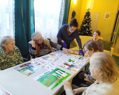 Przy siedzą stole seniorki. Na blacie rozłożone kolorowe plansze i obrazki. Obok terapeutka Magdalena Poraj-Górska.