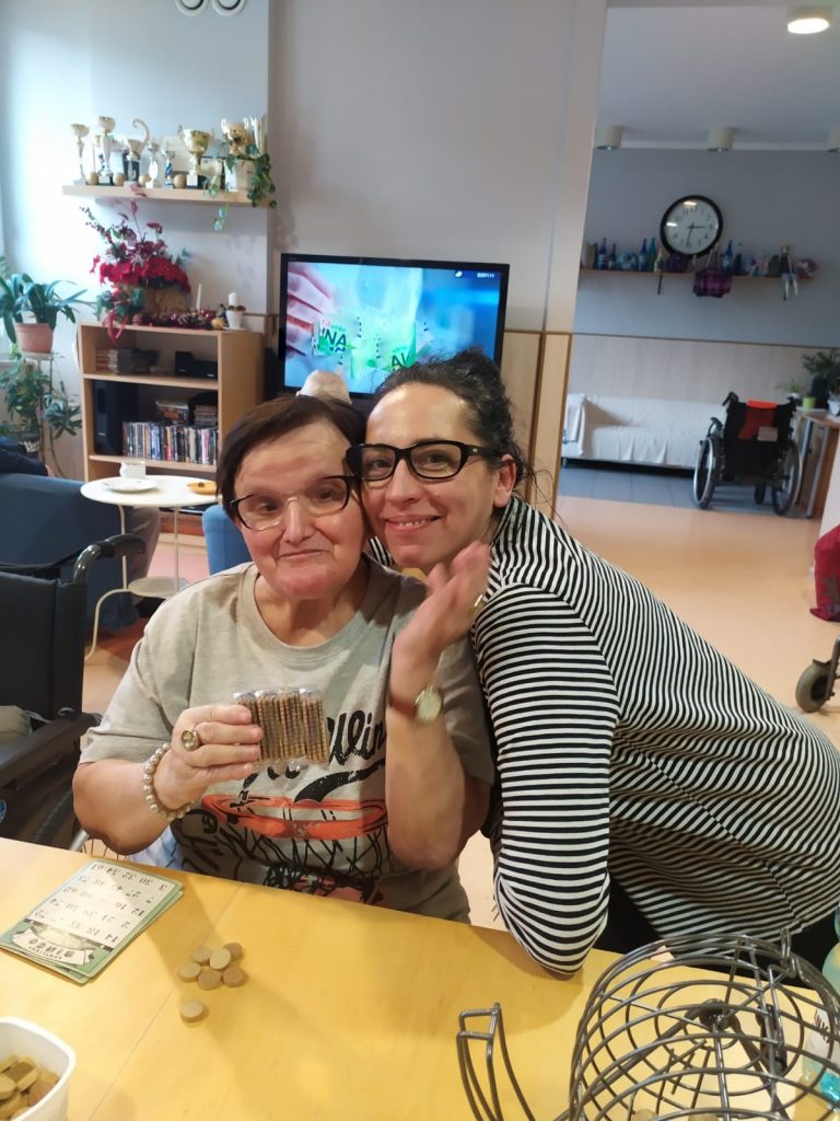 Świetlica. Terapeutka Magdalena Poraj-Górska przytula seniorkę. Uśmiechają się. Na stole gra Bingo.