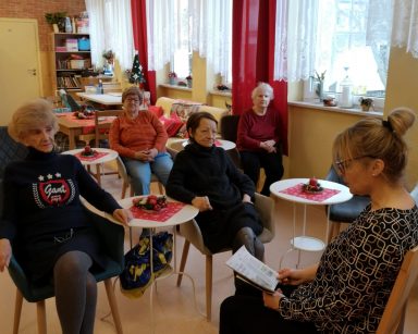 Sala. Terapeutka Beata Brzozowska siedzi na krześle, trzyma gazetę. Przed nią przy stolikach seniorzy.