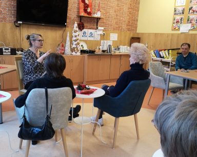 Sala. Terapeutka Beata Brzozowska siedzi na krześle. Przed nią przy stolikach seniorzy. Patrzą na nią, słuchają.
