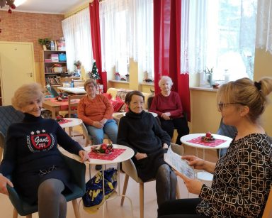 Sala. Terapeutka Beata Brzozowska siedzi na krześle. Przed nią przy stolikach seniorzy. Patrzą na nią, słuchają.