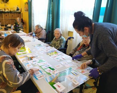 Przy siedzą stole seniorki. Na blacie rozłożone kolorowe plansze i obrazki. Obok terapeutka Magdalena Poraj-Górska.