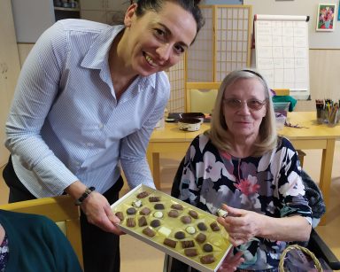 Świetlica. Terapeutka Magdalena Poraj-Górska częstuje seniorkę czekoladką z bombonierki. Uśmiechają się.