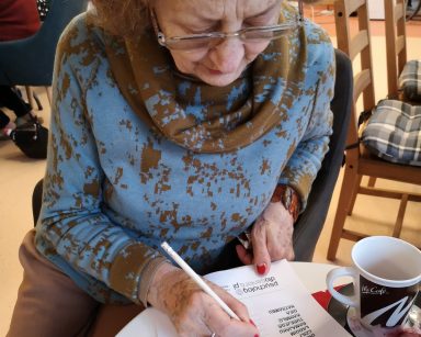 Seniorka siedzi przy stole. Nachyla się nad kartką i pisze. Obok na blacie kubek z kawą.