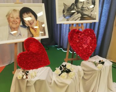 Na sali dekoracja. Na podeście biały materiał. Na nim białe róże, czerwone serca. Z tyłu dwa oprawione zdjęcia z seniorkami.