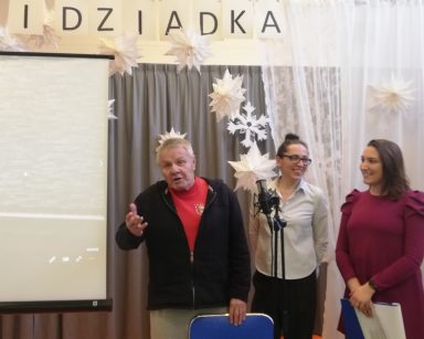 Sala. Przy mikrofonie stoi senior, fizjoterapeutka Martyna Józefczyk, terapeutka Magdalena Poraj-Górska.