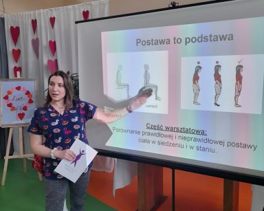 Sala gimnastyczna. Fizjoterapeutka Martyna Józefczyk przy ekranie projektora. Na nim slajd o prawidłowej postawie.