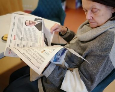 Sala. Seniorka siedzi na krześle. Czyta gazetę.