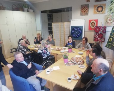 Świetlica. Dyrektorka Agnieszka Cysewska, pracownicy i seniorzy siedzą przy faworkach, kawie i herbacie. Jedzą, rozmawiają.