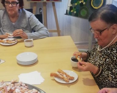 Świetlica. Seniorki siedzą przy stole. Jedzą faworki. Przed nimi talerze z faworkami, kubki z kawą.