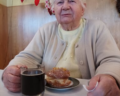 Sala. Seniorka siedzi przy stoliku. Uśmiecha się, trzyma kubek z kawą. Na blacie talerz z pączkiem.