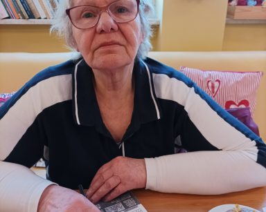 Sala. Seniorka siedzi przy stoliku. Na blacie talerz z pączkiem, krzyżówka.